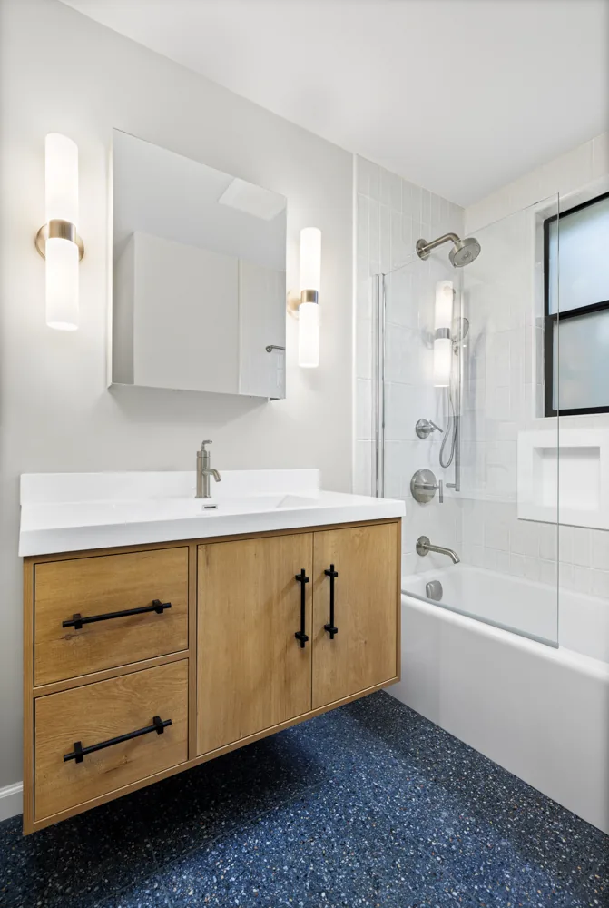 A full master bathroom remodel featuring a custom floating wood vanity, white subway tile, a tub-and-shower combination, and a striking speckled terrazzo floor.